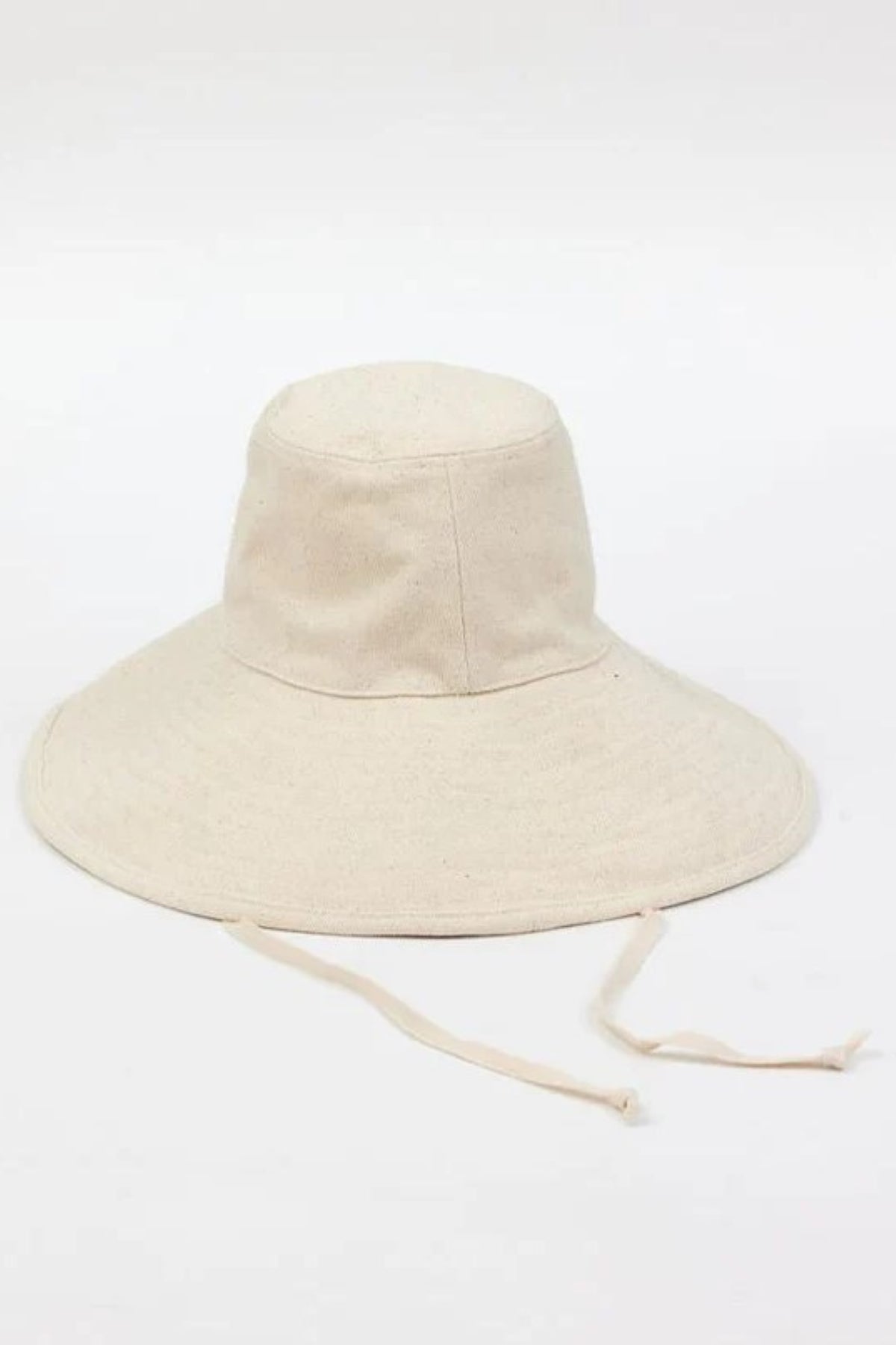 The LIVVY CANVAS SUN HAT from Velvet by Graham & Spencer, a wide-brimmed light beige fabric hat with adjustable strings, rests on a plain white surface.-38895651061953
