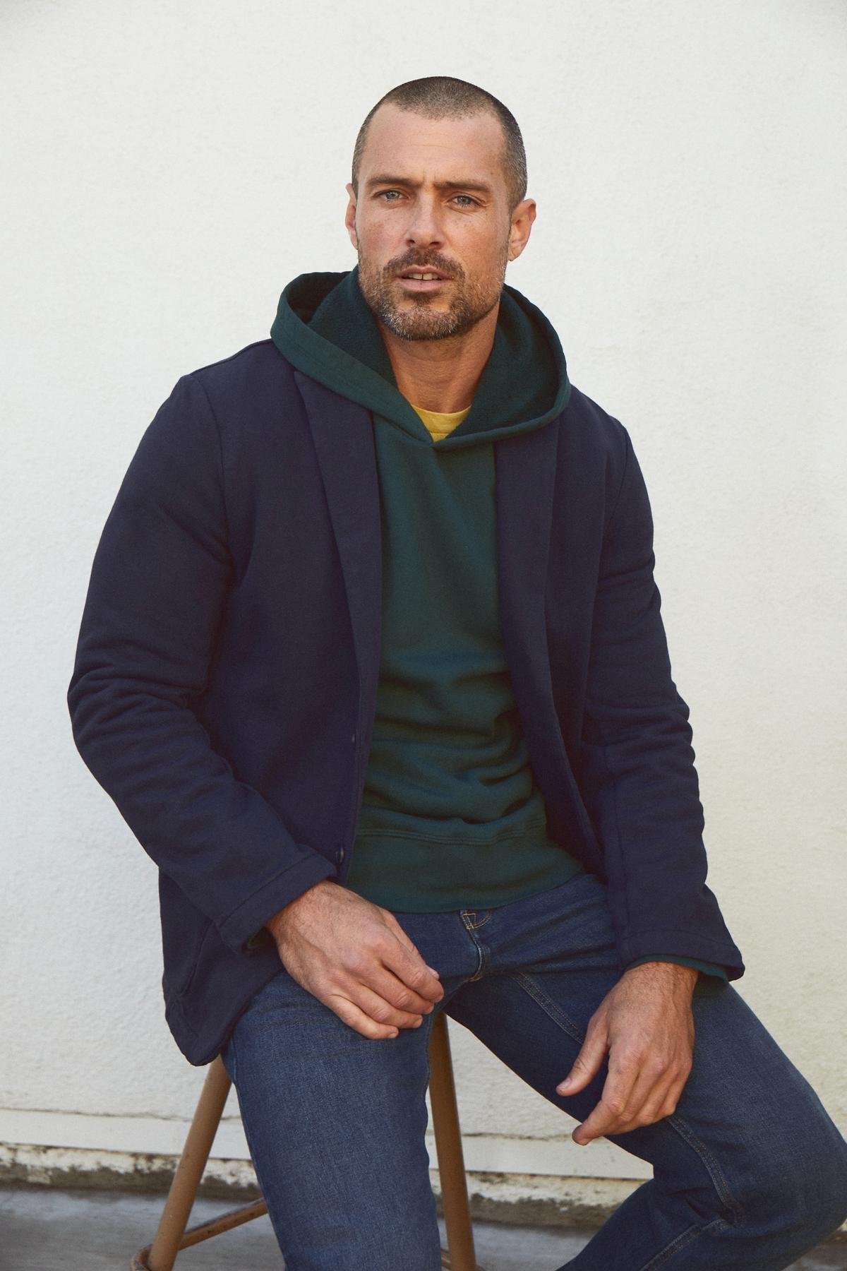 A man wearing a Velvet by Graham & Spencer HAYDEN STRUCTURED BLAZER and jeans sits on a chair.-35231587664065