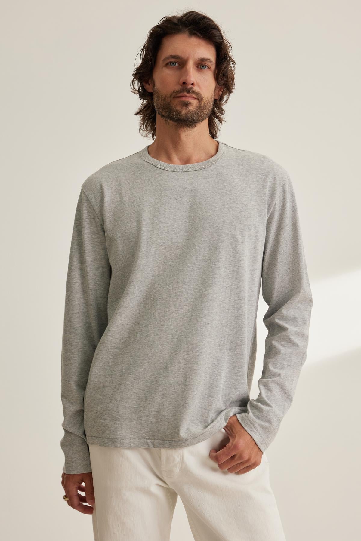 A man with long hair and a beard wears the EVREN TEE by Velvet by Graham & Spencer, paired with white pants, against a neutral background.