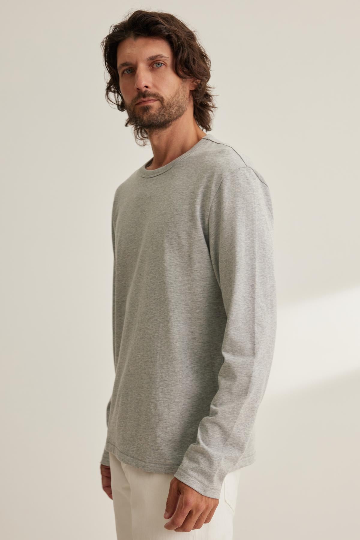 A man with long hair and a beard models the EVREN TEE in light gray by Velvet by Graham & Spencer, paired with white pants, against a plain background.