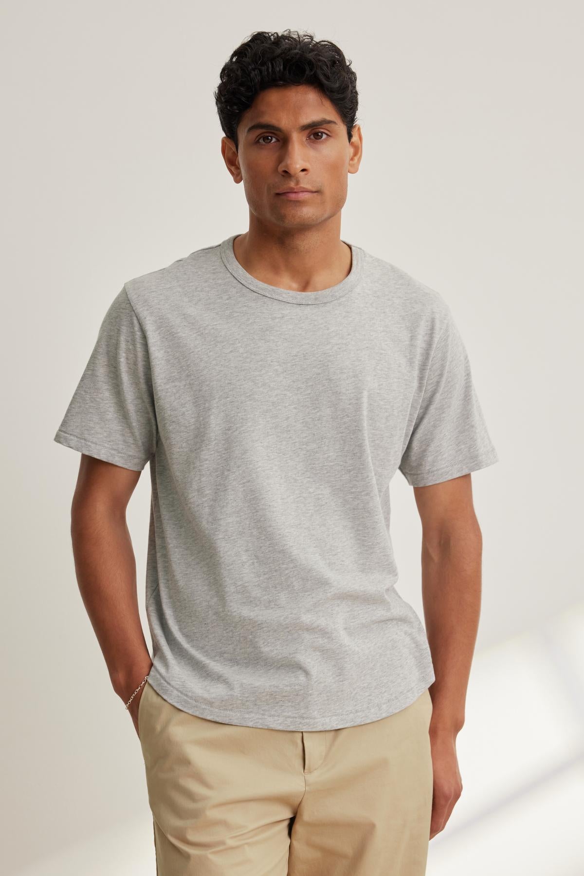 A man wearing the MADDEN TEE by Velvet by Graham & Spencer and beige pants stands with one hand in his pocket against a plain background.