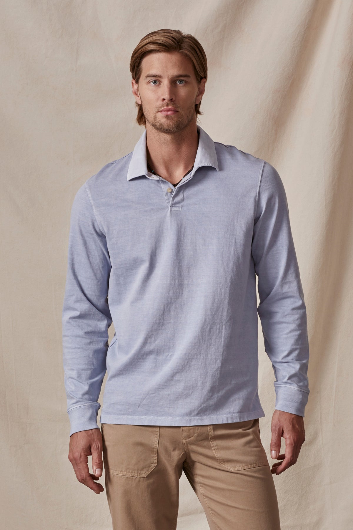 A man wearing a light blue long-sleeve BALTHAZAR POLO by Velvet by Graham & Spencer and beige pants stands in front of a beige fabric backdrop.-36009337487553