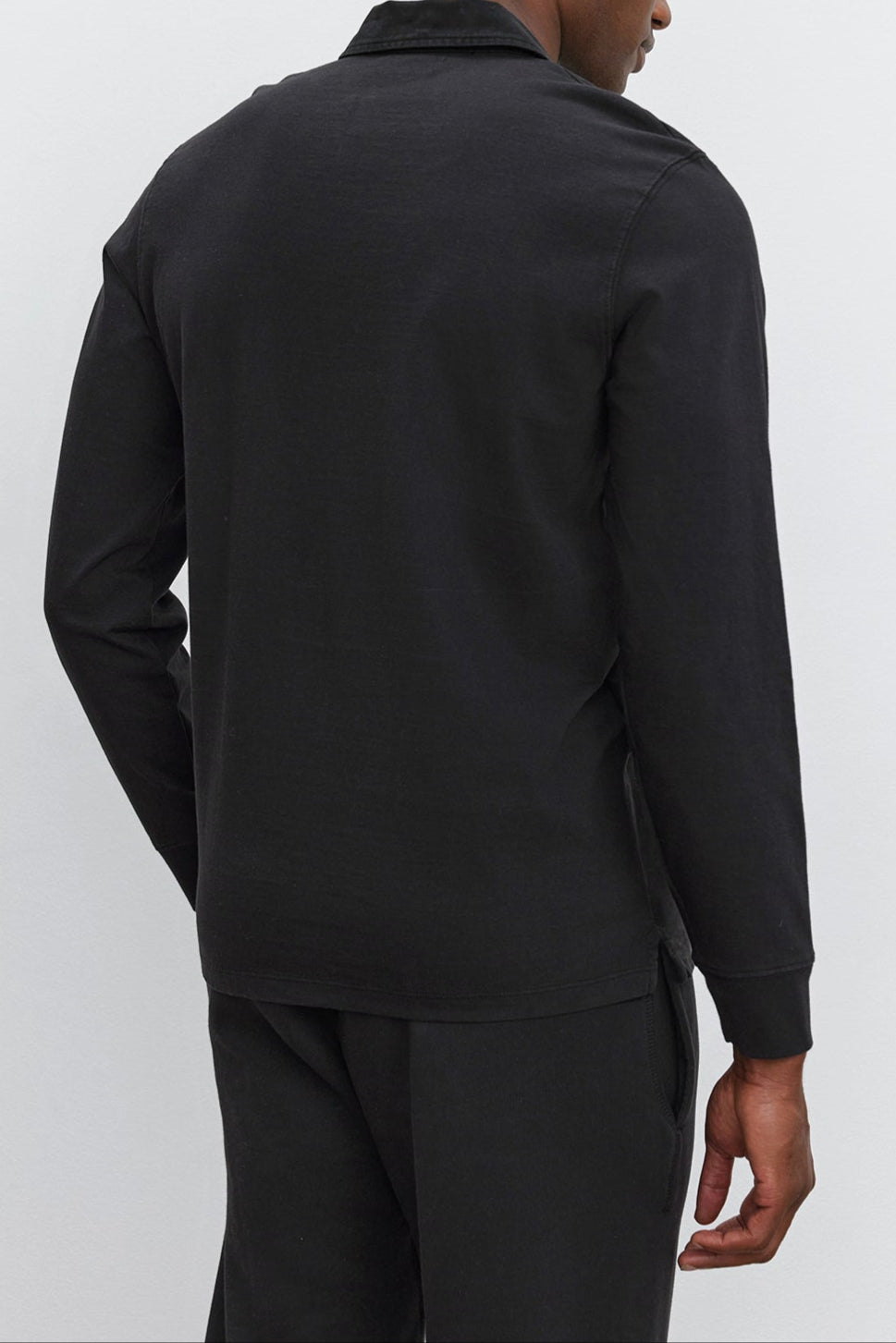 A person is seen from the back, standing against a plain, light-colored background. They are wearing a long-sleeve black BALTHAZAR POLO by Velvet by Graham & Spencer along with black pants—a versatile addition to any wardrobe.-37893747540161
