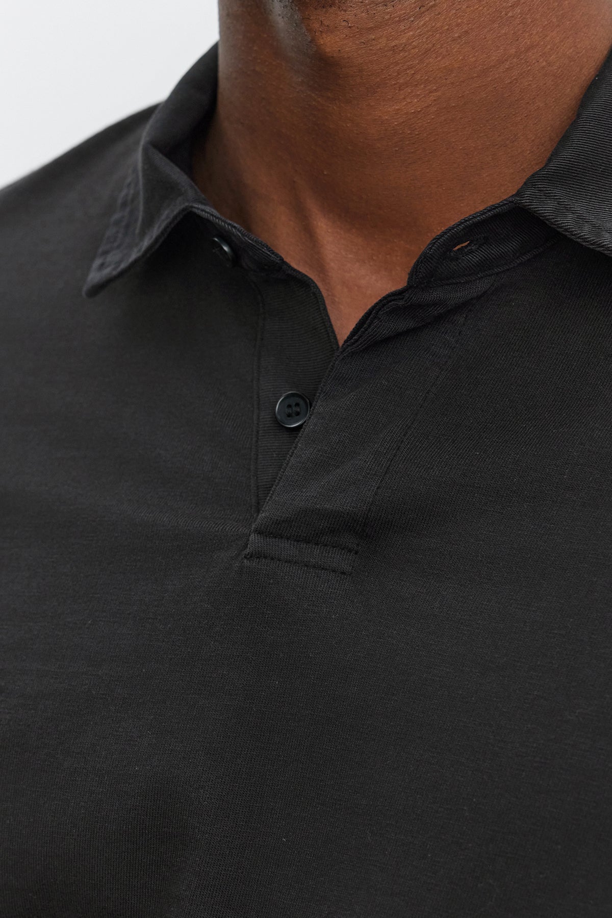 Close-up of a person wearing the BALTHAZAR POLO by Velvet by Graham & Spencer, a black rugby-style polo crafted from garment-dyed cotton jersey with a partially buttoned placket. Only the neck and upper chest area are visible.-37893747704001