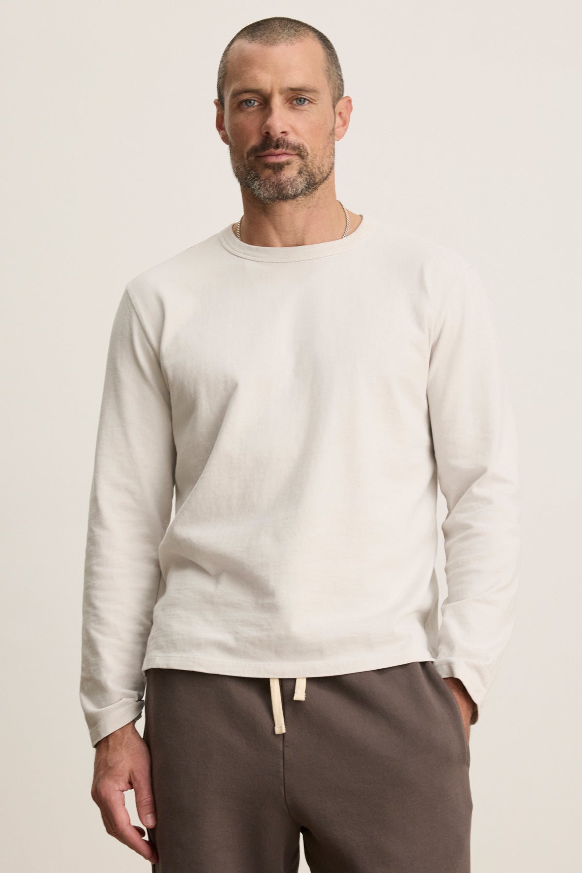 A man with short hair and a beard wears the BRADY TEE by Velvet by Graham & Spencer, an off-white midweight jersey long-sleeve shirt, paired with brown drawstring pants, perfect for everyday wear against a neutral background.