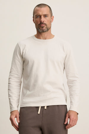 A man with short hair and a beard wears the Velvet by Graham & Spencer BRADY TEE, a plain white midweight jersey long-sleeve shirt, paired with brown drawstring pants, standing against a neutral background.