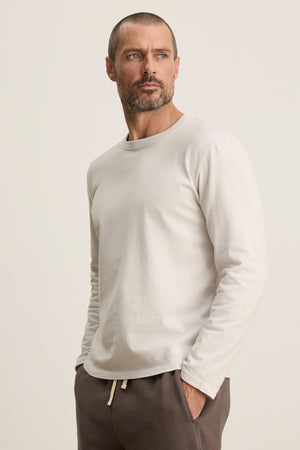 A man with short hair and a beard wears the Velvet by Graham & Spencer BRADY TEE, a light long-sleeve midweight jersey shirt, paired with tailored dark pants—an effortless everyday look standing against a plain background.