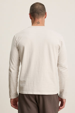A man with a short haircut and a bird tattoo on his neck is seen from behind wearing the Velvet by Graham & Spencer BRADY TEE, a tailored fit, long-sleeve off-white midweight jersey shirt, with brown pants against a plain background.