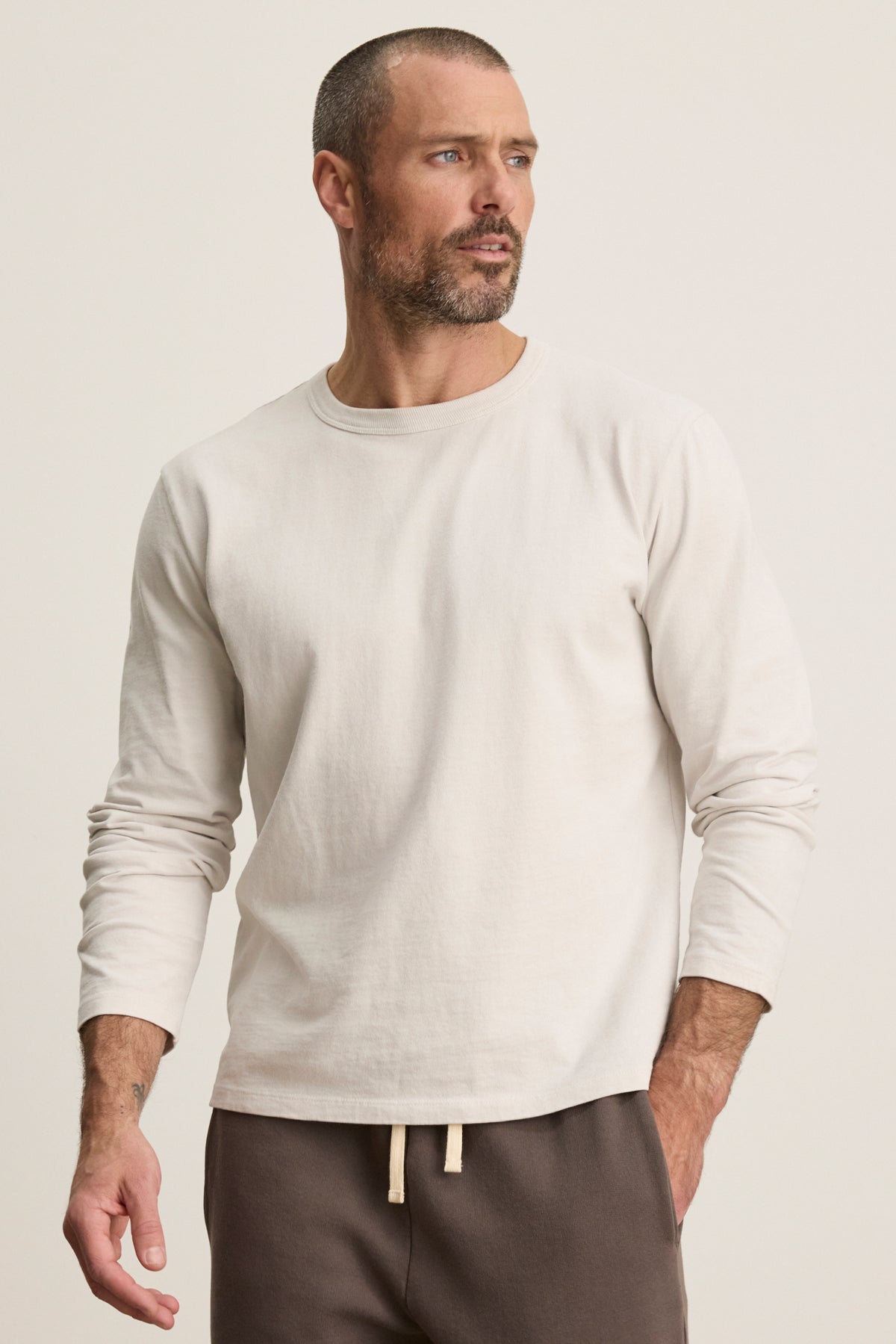 A man wears the BRADY TEE by Velvet by Graham & Spencer—a light beige, midweight jersey long-sleeve—paired with brown drawstring pants against a plain background, offering an effortlessly refined everyday look.