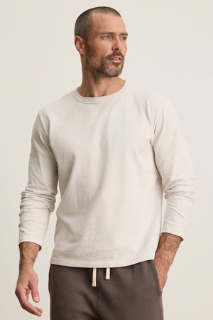 A man wears the BRADY TEE by Velvet by Graham & Spencer—a light beige, midweight jersey long-sleeve—paired with brown drawstring pants against a plain background, offering an effortlessly refined everyday look.