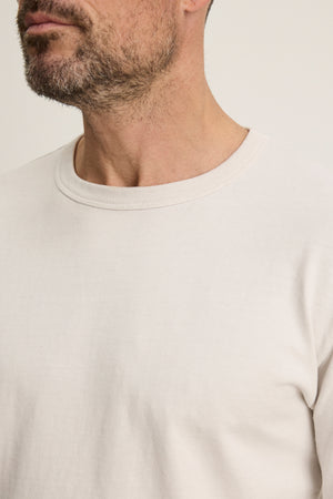 A man with a short beard and mustache wears the BRADY TEE by Velvet by Graham & Spencer, an off-white, tailored fit crew neck shirt in midweight jersey, ideal for everyday wear, set against a plain background.