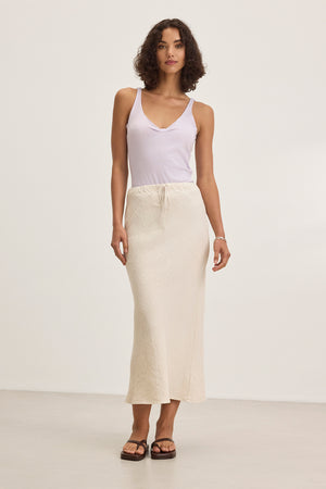 A person with curly hair models a white tank top, the BEKA HEAVY LINEN SKIRT by Velvet by Graham & Spencer in cream, and black sandals against a plain white background.