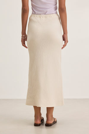Back view of a person wearing a white top and the BEKA HEAVY LINEN SKIRT by Velvet by Graham & Spencer in beige, bias cut maxi style, paired with black sandals, standing against a plain background.