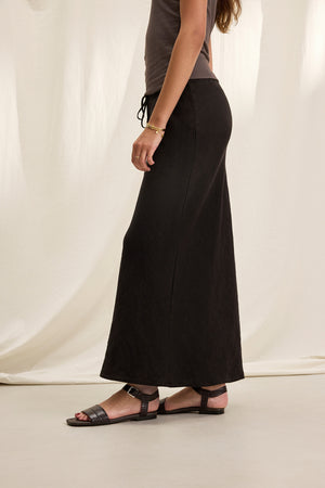 Side view of a person wearing a gray top, the BEKA HEAVY LINEN SKIRT by Velvet by Graham & Spencer, and black sandals, standing against a light curtain background.
