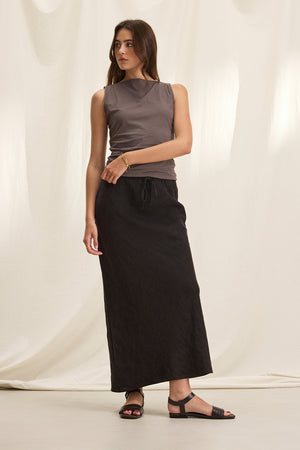 A woman stands against a white backdrop, wearing a sleeveless gray top, the BEKA HEAVY LINEN SKIRT by Velvet by Graham & Spencer with a graceful bias cut, and black sandals.