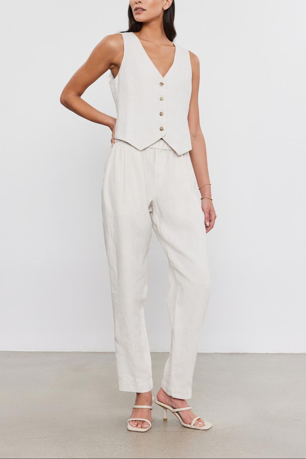 A woman stands in a studio, wearing a white sleeveless top with buttons, paired with matching Jessie Heavy Linen pants from Velvet by Graham & Spencer and light sandals.-36910040875201