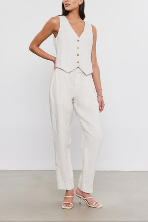 A woman stands in a studio, wearing a white sleeveless top with buttons, paired with matching Jessie Heavy Linen pants from Velvet by Graham & Spencer and light sandals.
