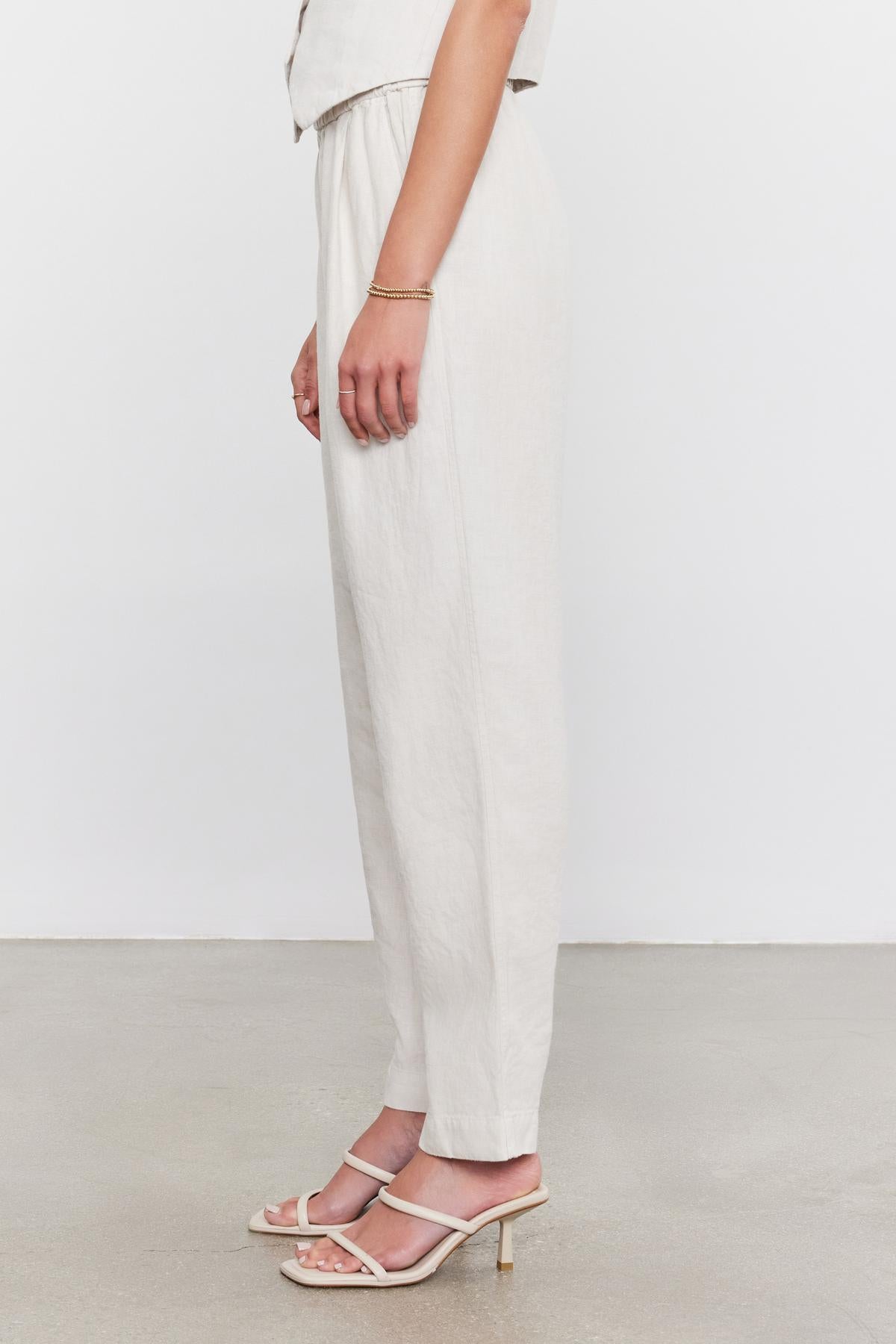 Woman standing in a studio, cropped at the waist, wearing white Velvet by Graham & Spencer Jessie Heavy Linen pants and heeled sandals, with a hand slightly touching her side.-36910040907969