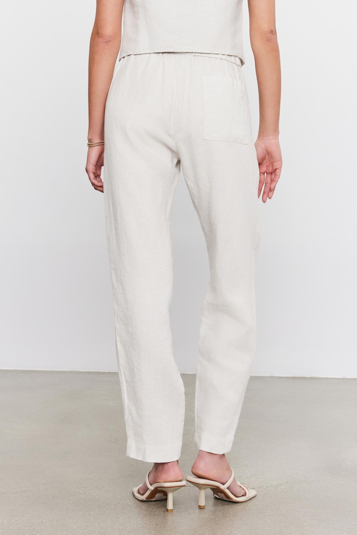 Rear view of a person wearing Velvet by Graham & Spencer cream linen trousers and tan slingback heels, standing against a white background.-36910040940737