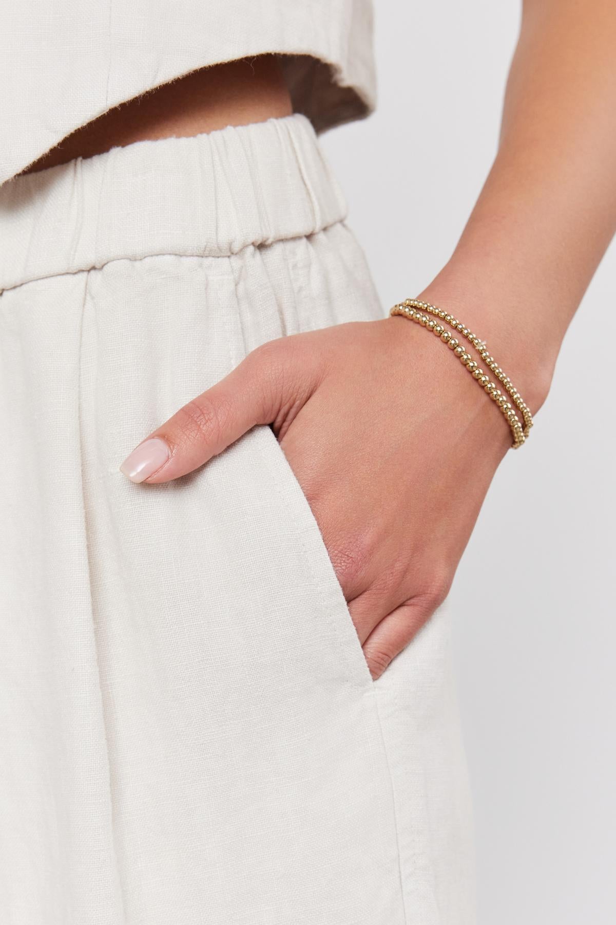 Woman's hand with pink manicure resting in the pocket of her beige Velvet by Graham & Spencer JESSIE HEAVY LINEN PANT trousers, wearing a gold bracelet.-36910040973505