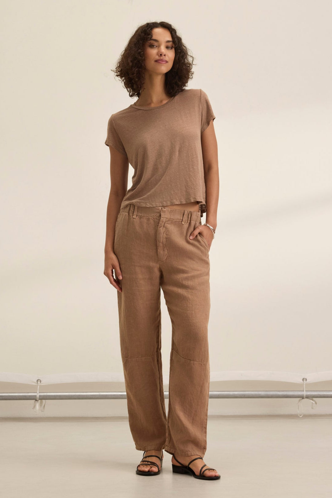 A woman with curly hair wears a neutral-toned outfit, featuring Velvet by Graham & Spencer's tailored TAYLOR HEAVY LINEN PANT in brown and a matching t-shirt, completed with black sandals against a light background.-38821051793601