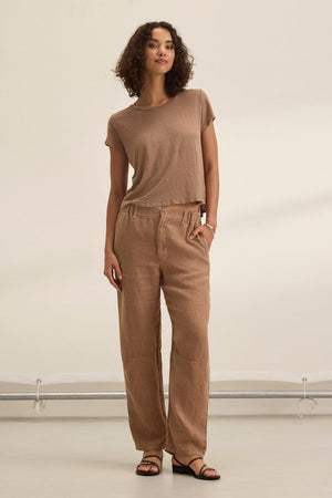 A woman with curly hair wears a neutral-toned outfit, featuring Velvet by Graham & Spencer's tailored TAYLOR HEAVY LINEN PANT in brown and a matching t-shirt, completed with black sandals against a light background.