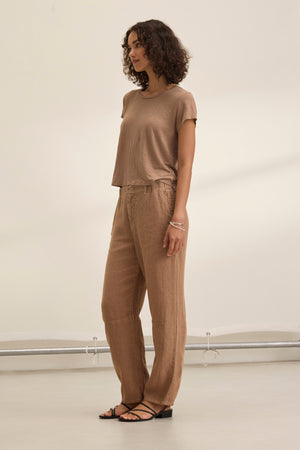 A person with curly hair wears the tailored TAYLOR HEAVY LINEN PANT by Velvet by Graham & Spencer in a neutral tone, paired with a short-sleeved top, against a plain background.