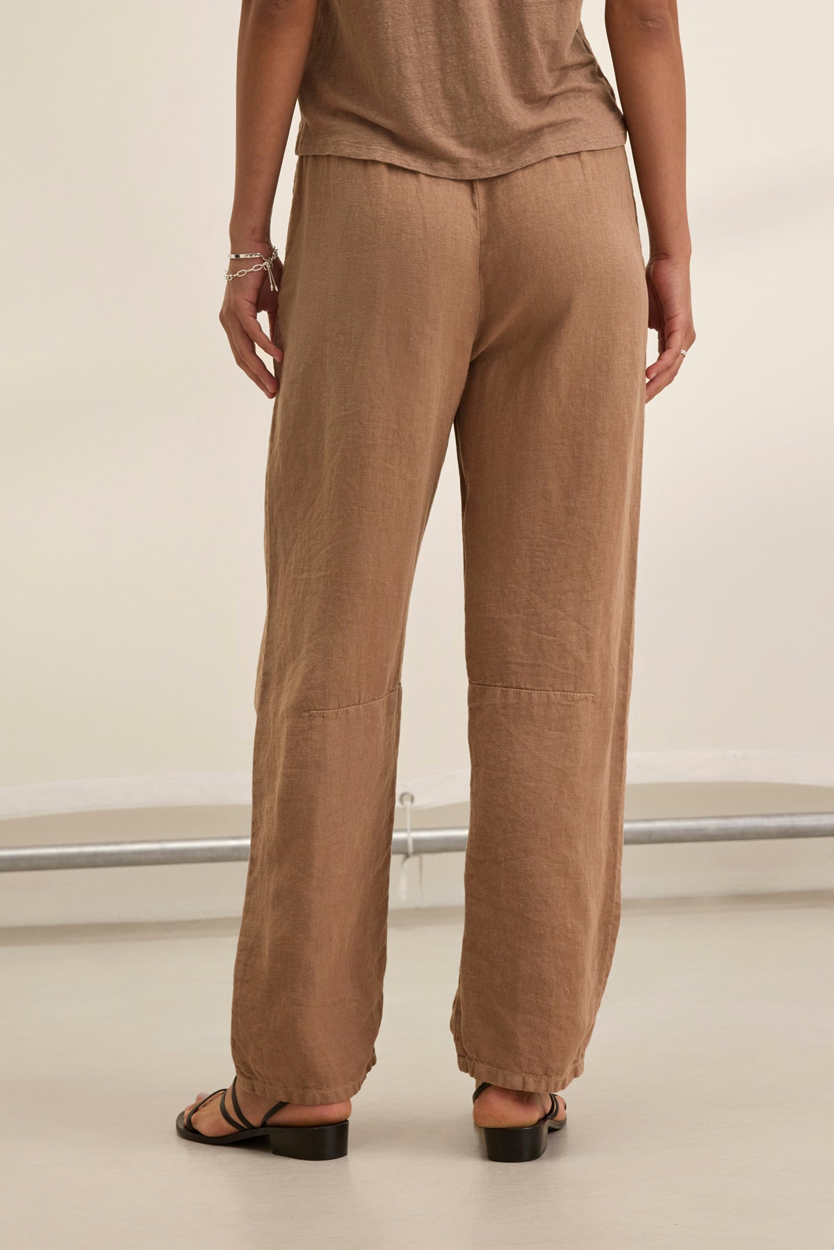 Someone wearing Velvet by Graham & Spencer's TAYLOR HEAVY LINEN PANT in brown and flat sandals stands on a light-colored floor, facing away.-38821051859137