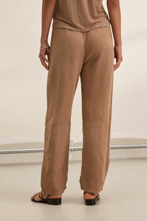 Someone wearing Velvet by Graham & Spencer's TAYLOR HEAVY LINEN PANT in brown and flat sandals stands on a light-colored floor, facing away.