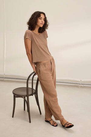 A person with curly hair, dressed in a Velvet by Graham & Spencer's TAYLOR HEAVY LINEN PANT and a brown t-shirt, leans casually against a wooden chair in a minimalist setting.