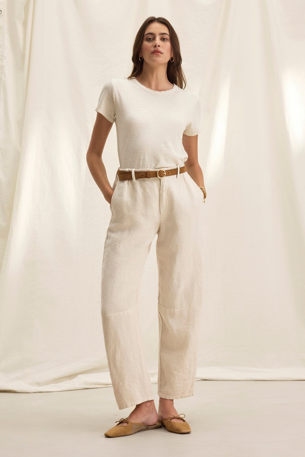 A woman in a white t-shirt and tailored fit TAYLOR HEAVY LINEN PANT by Velvet by Graham & Spencer, accessorized with a brown belt and tan flats, stands confidently with her hands in pockets against a light backdrop.-38821052088513