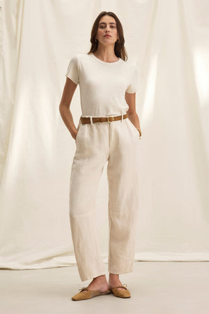 A woman in a white t-shirt and tailored fit TAYLOR HEAVY LINEN PANT by Velvet by Graham & Spencer, accessorized with a brown belt and tan flats, stands confidently with her hands in pockets against a light backdrop.