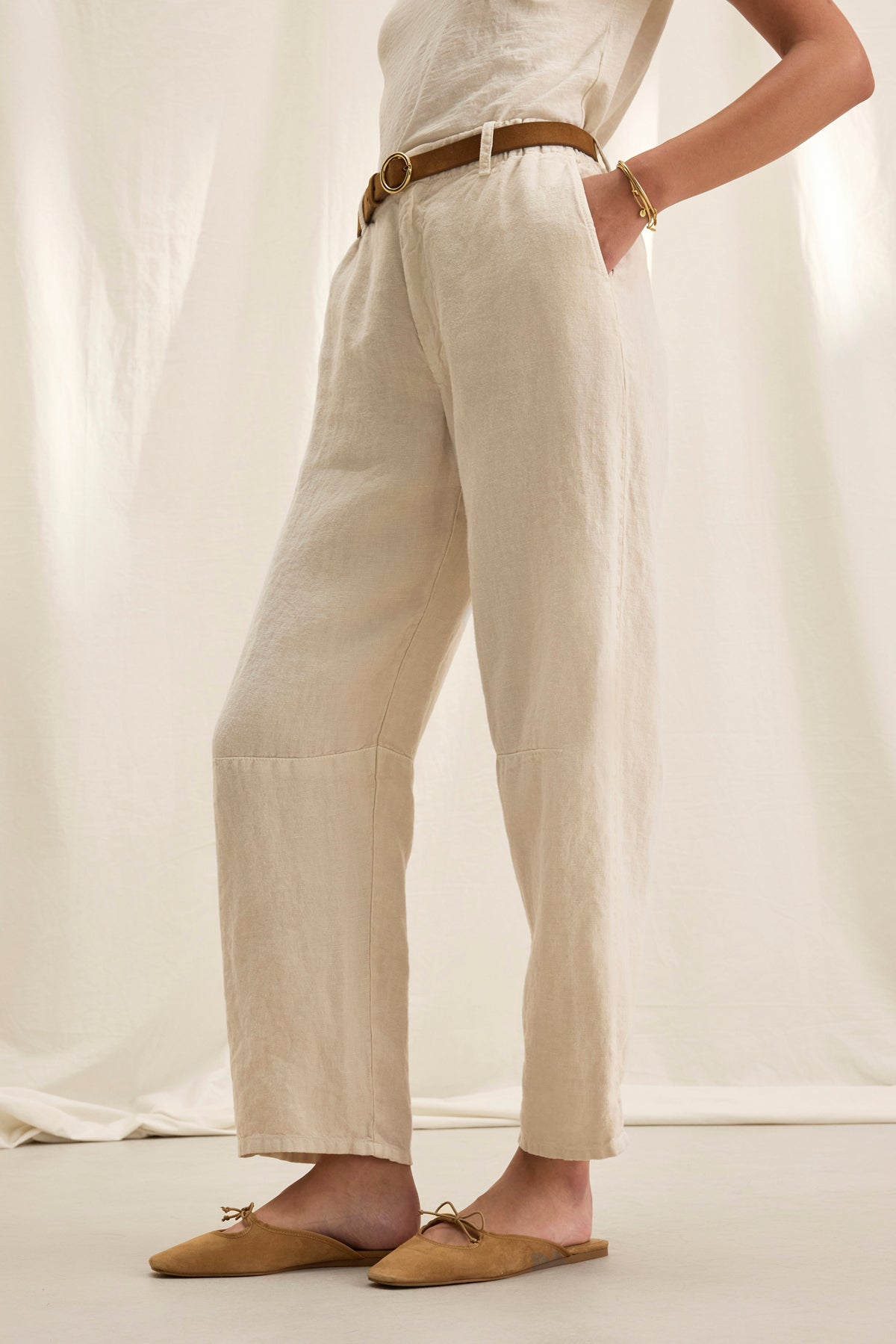 Wearing Velvet by Graham & Spencer's TAYLOR HEAVY LINEN PANT, a person dons a chic ensemble with tailored beige pants and top, accented by a structured silhouette. A brown belt and slippers finish the look against a neutral background.-38821052154049