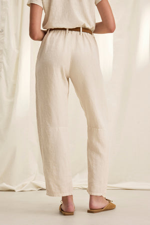 A person wearing TAYLOR HEAVY LINEN PANT from Velvet by Graham & Spencer, along with a matching top, stands against a beige draped fabric background, highlighting the tailored fit and structured silhouette.