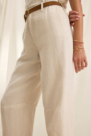 A person in tailored TAYLOR HEAVY LINEN PANT by Velvet by Graham & Spencer paired with a brown belt. One arm rests on their hip, adorned with two gold bracelets adding elegance to the structured silhouette.