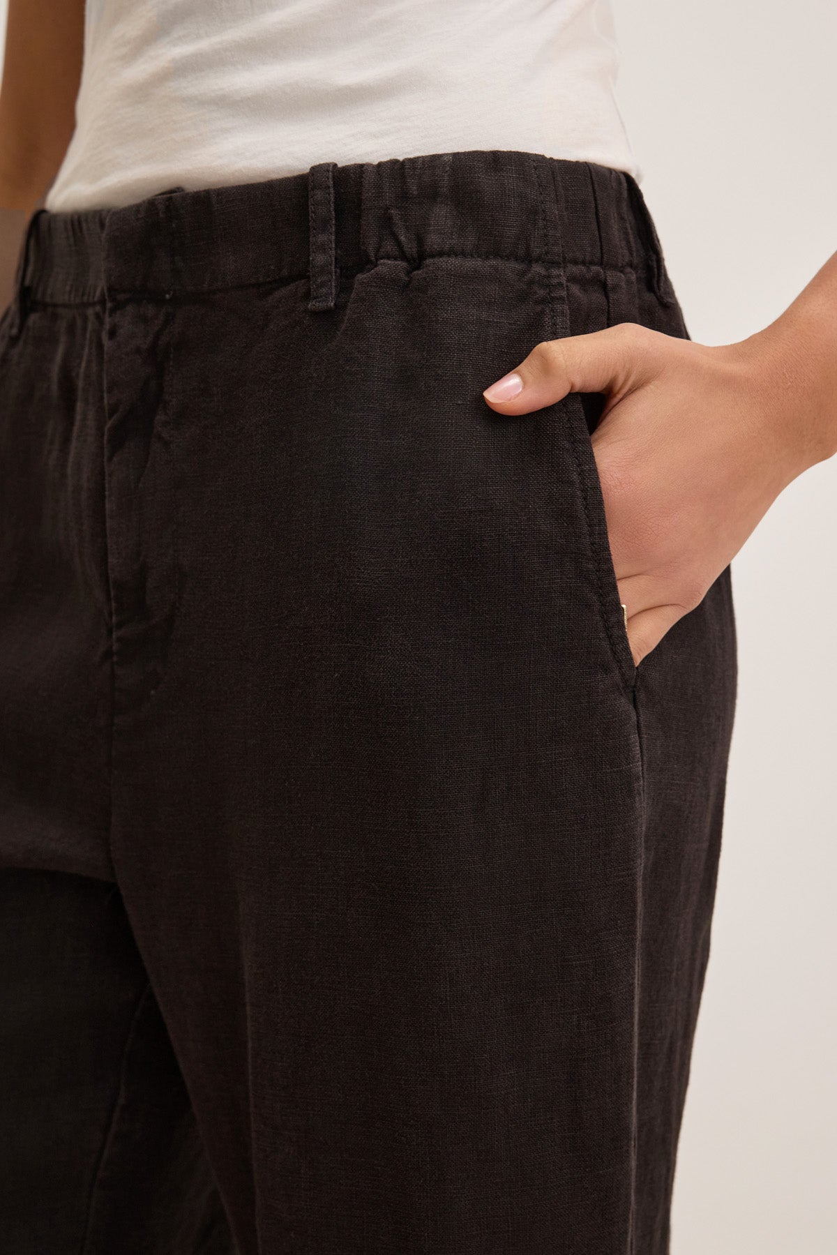 A person wearing Velvet by Graham & Spencer’s TAYLOR HEAVY LINEN PANT in black, featuring a tailored fit and curved leg silhouette, paired with a white shirt, stands with one hand in their pocket.-41235234914497