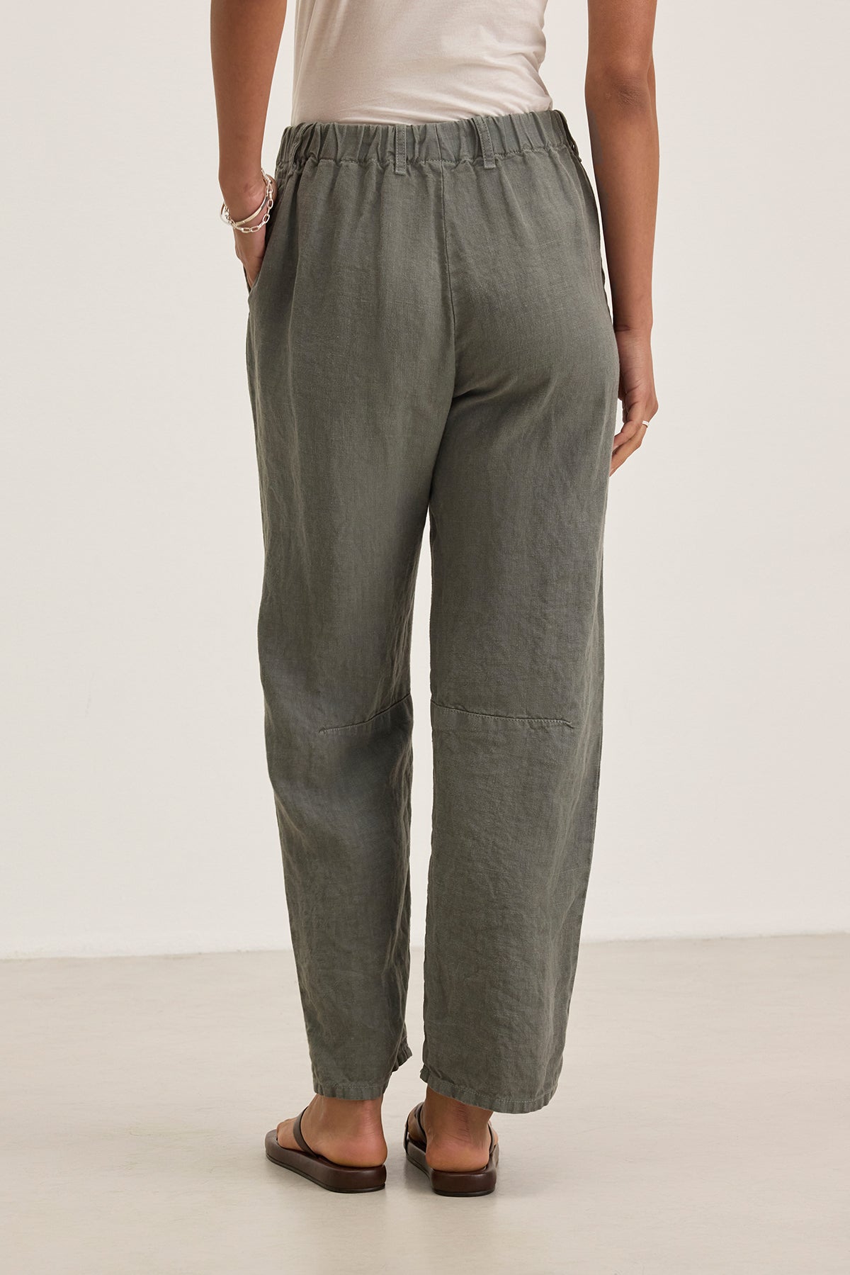 A person wearing the TAYLOR HEAVY LINEN PANT from Velvet by Graham & Spencer in olive green, tailored and loose-fitting, stands with hands in pockets, showcasing the back view paired with a light top.