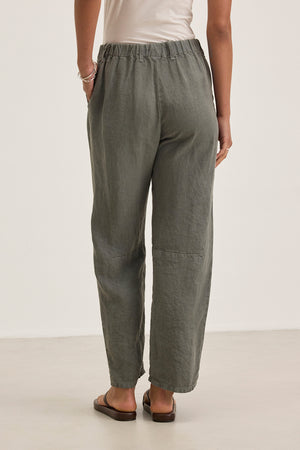 A person wearing the TAYLOR HEAVY LINEN PANT from Velvet by Graham & Spencer in olive green, tailored and loose-fitting, stands with hands in pockets, showcasing the back view paired with a light top.