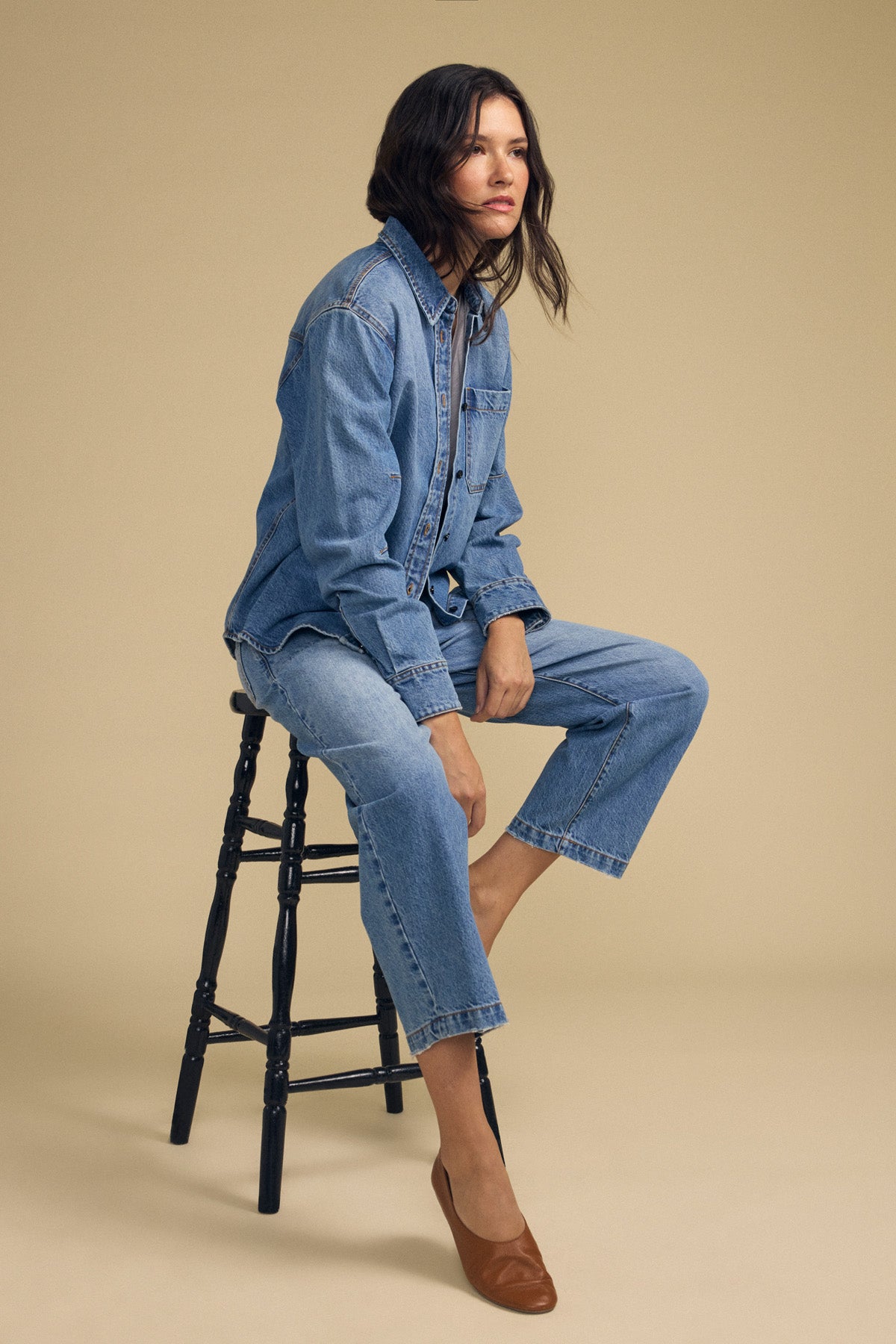 A woman wearing the CLEMENTINE DENIM SHIRT by Velvet by Graham & Spencer and jeans sits on a wooden stool against a plain beige background, looking to the side.-41814471475393