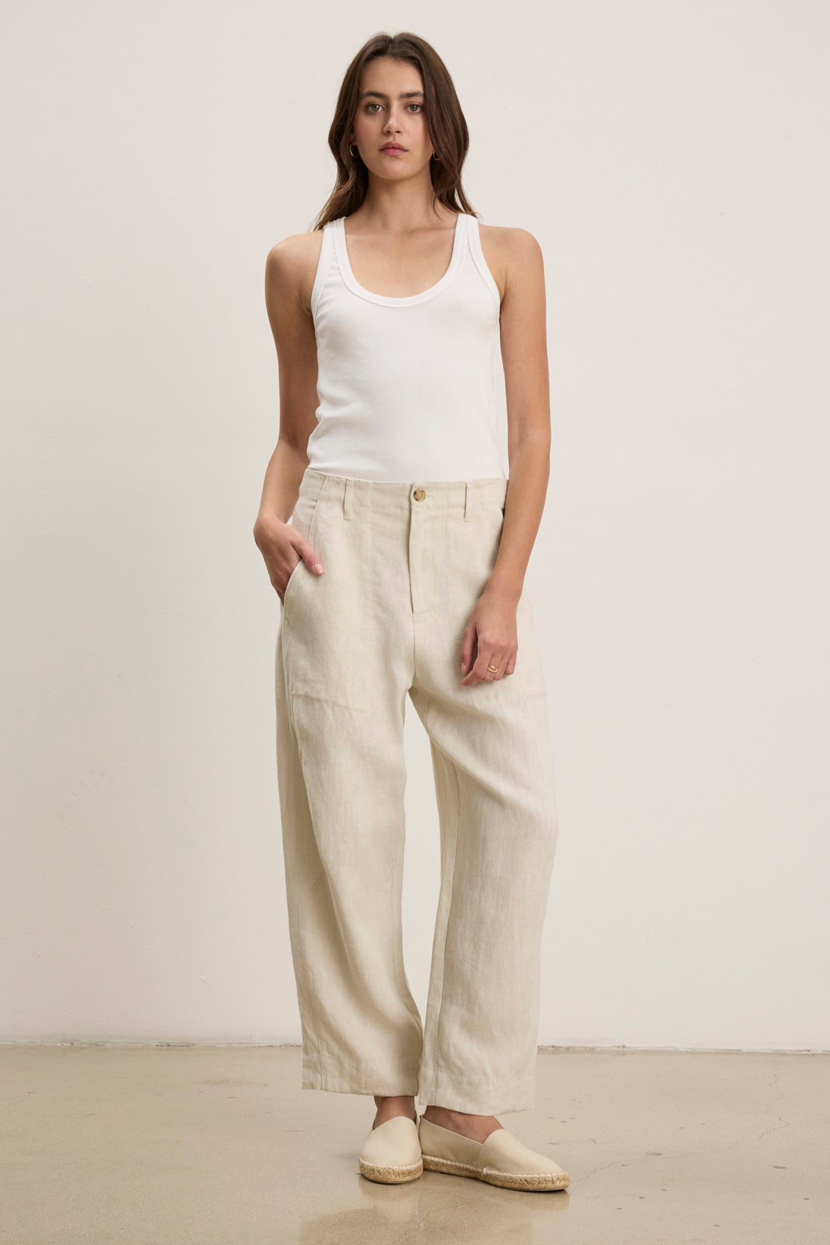 A woman stands against a plain background wearing a white tank top, ISSA HEAVY LINEN PANT by Velvet by Graham & Spencer in beige with a relaxed silhouette, and beige slip-on shoes.
