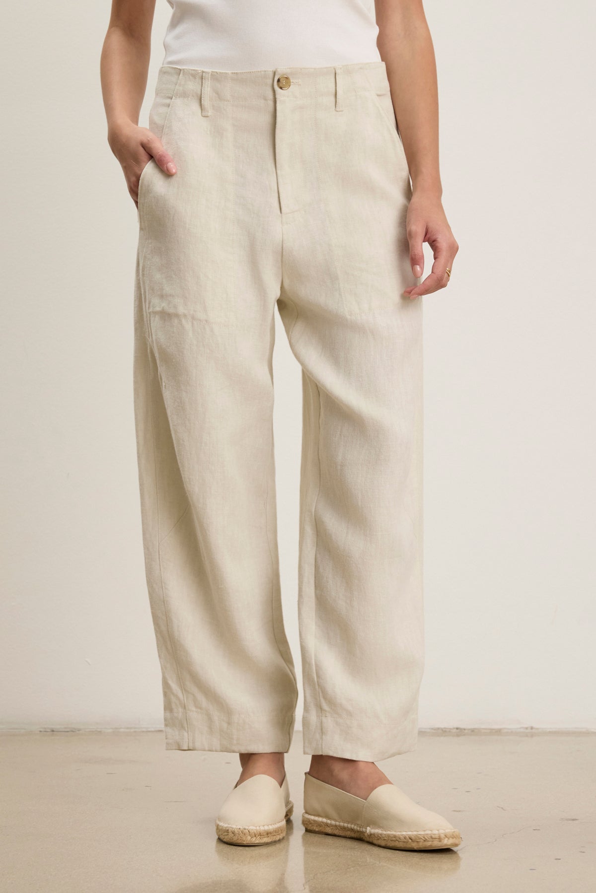 A person stands against a plain background wearing Velvet by Graham & Spencer’s ISSA HEAVY LINEN PANT in light beige, paired with a white top and beige espadrilles, showing a relaxed silhouette with one hand in their pocket.