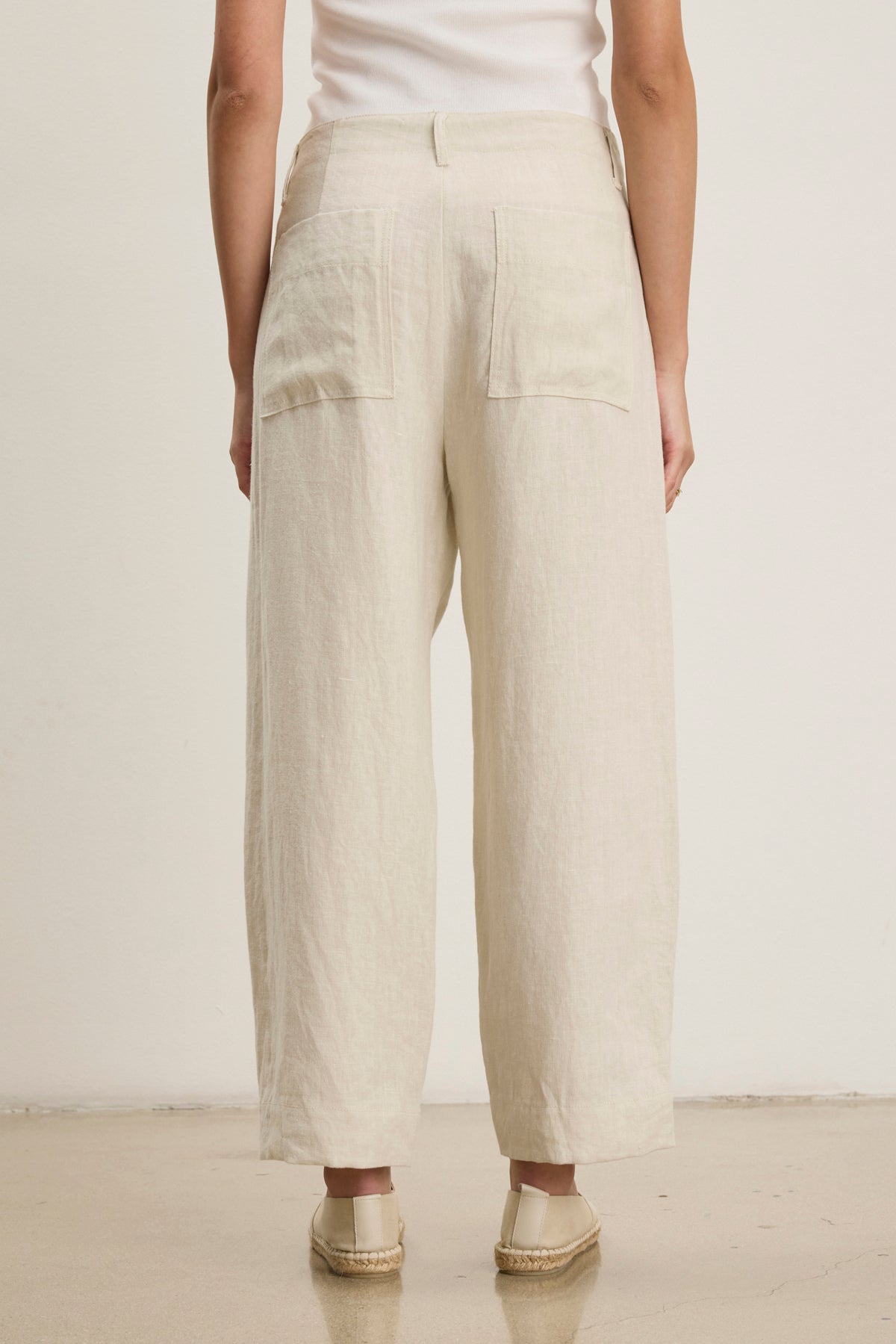 A person wearing Velvet by Graham & Spencer's ISSA HEAVY LINEN PANT in light beige, styled with a white top and beige espadrilles, stands on a light-colored floor facing away from the camera.