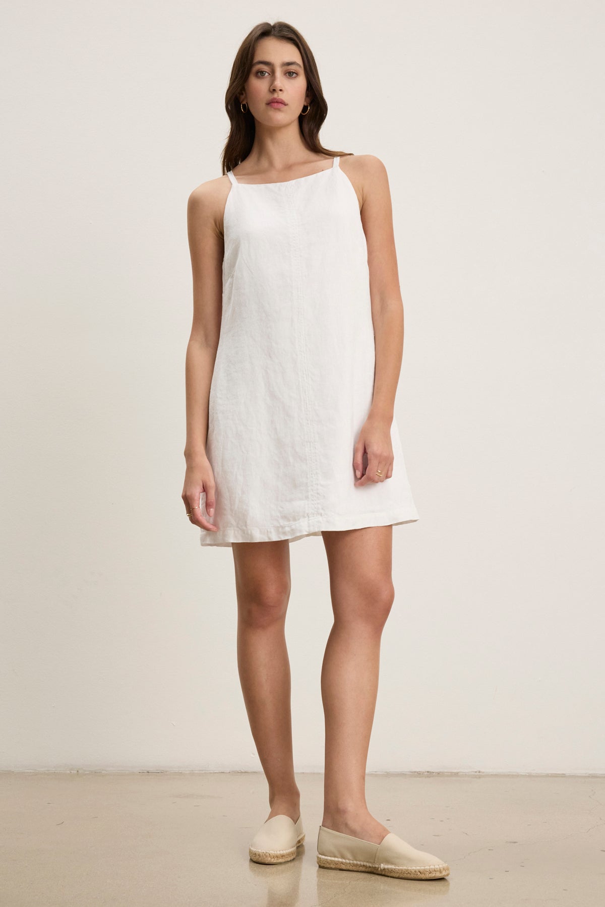 A woman stands against a plain background, wearing the NARA LINEN DRESS by Velvet by Graham & Spencer—a sleeveless, heavy linen mini dress with a square neckline—paired with beige espadrilles.