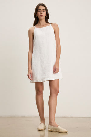 A woman stands against a plain background, wearing the NARA LINEN DRESS by Velvet by Graham & Spencer—a sleeveless, heavy linen mini dress with a square neckline—paired with beige espadrilles.