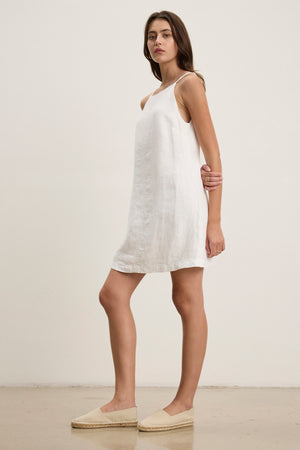 A woman stands sideways in the NARA LINEN DRESS by Velvet by Graham & Spencer—a sleeveless heavy linen mini dress with a square neckline—paired with beige espadrilles against a plain, light background.