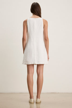 A woman faces away, wearing the NARA LINEN DRESS by Velvet by Graham & Spencer—a sleeveless mini dress with a square neckline—paired with light espadrilles, against a plain light background.
