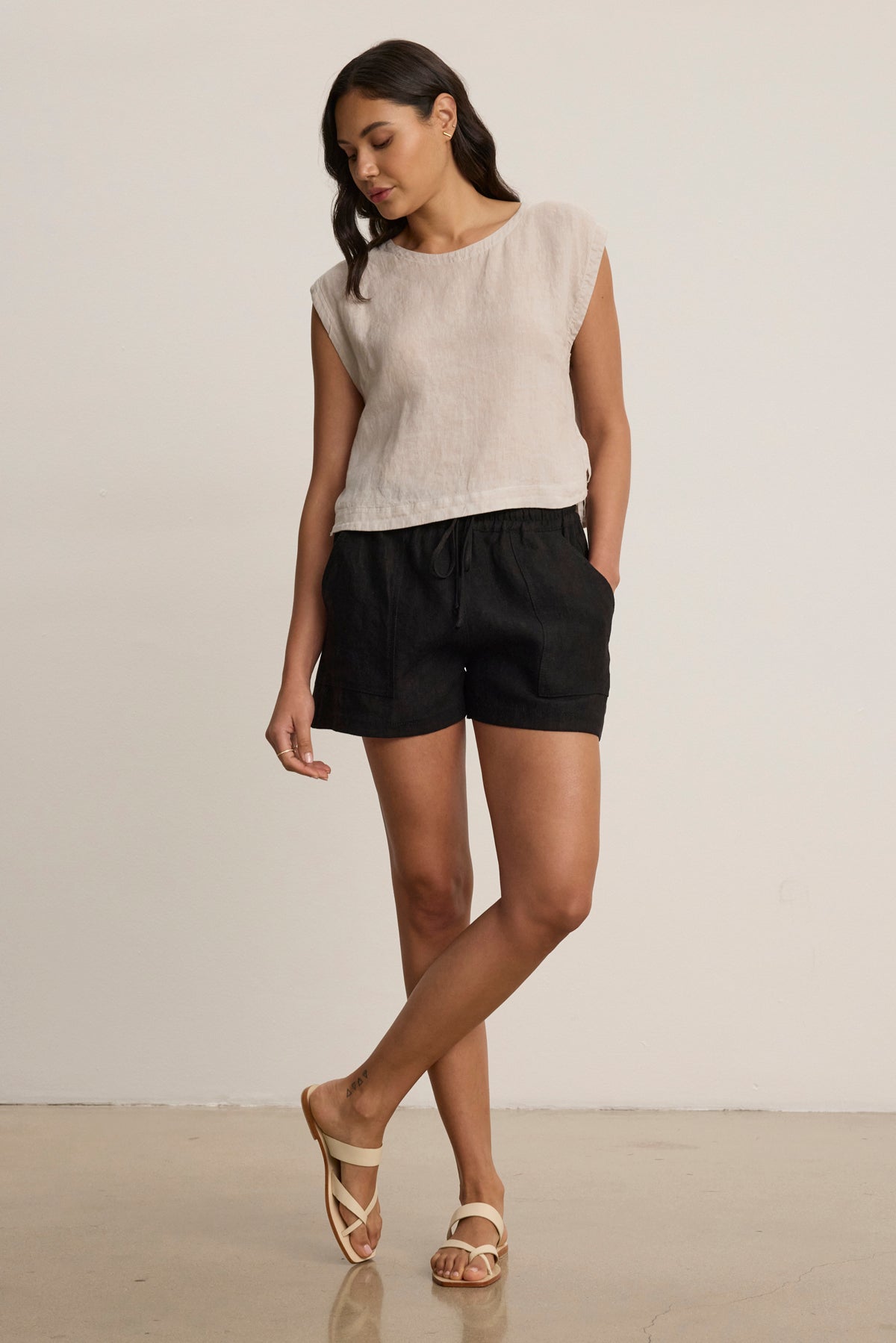 A woman stands indoors wearing a short-sleeved beige top, beige sandals, and the SUTTON HEAVY LINEN SHORT by Velvet by Graham & Spencer—black linen shorts with an elastic drawstring waist—crossing one leg in front of the other.-39123355664577