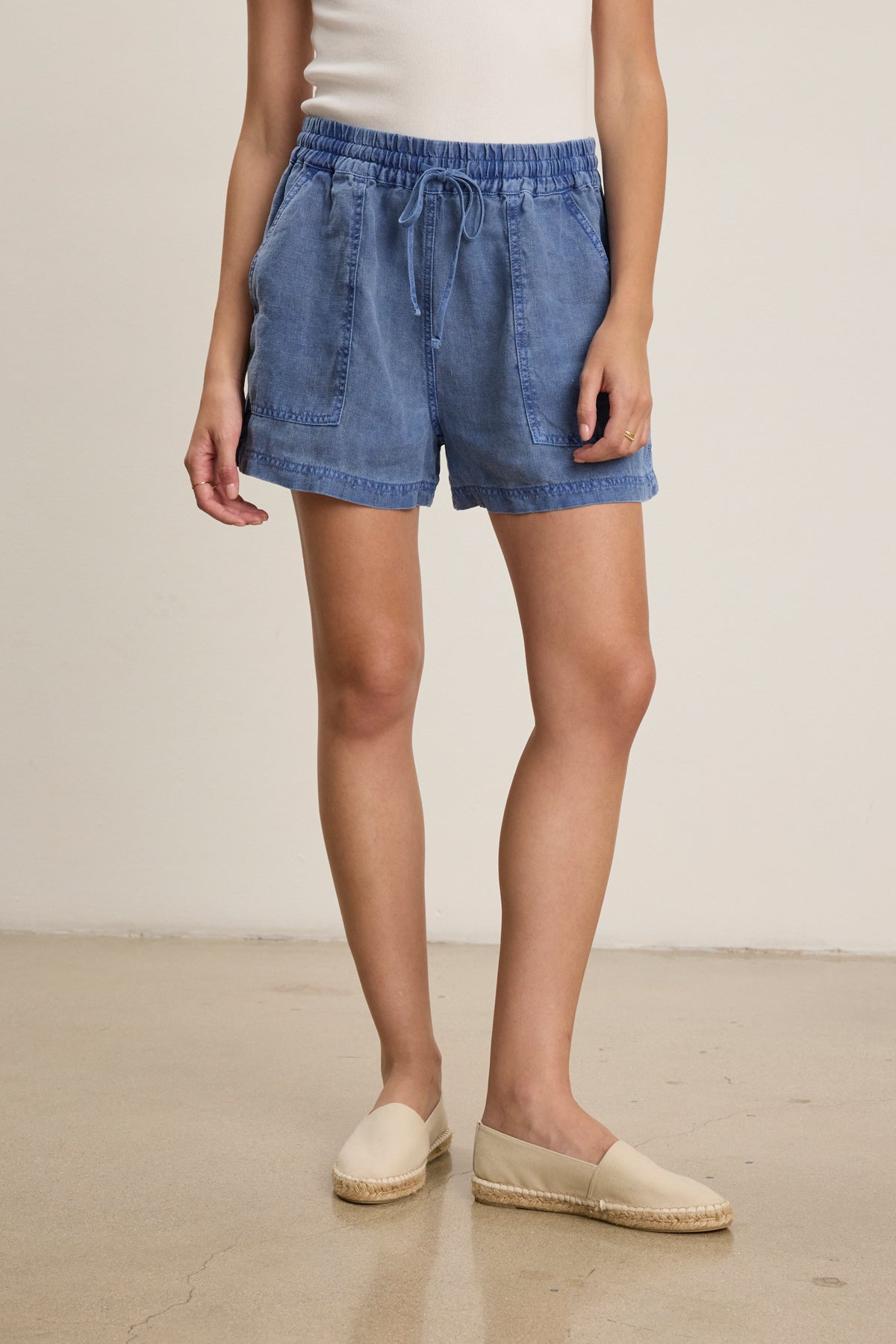 A person wears the SUTTON HEAVY LINEN SHORT by Velvet by Graham & Spencer—blue with an elastic drawstring waist—paired with a white sleeveless top and beige espadrilles, standing on a beige floor against a plain light background.