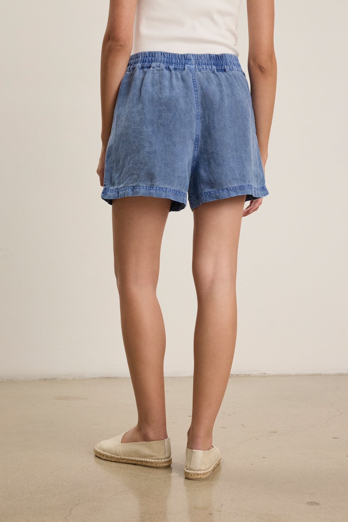 Wearing a white top, Velvet by Graham & Spencer's SUTTON HEAVY LINEN SHORT with an elastic drawstring waist, and beige espadrilles, a person stands indoors on a polished floor facing away from the camera.