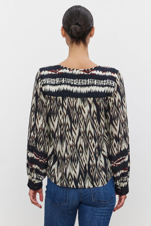 A person with dark hair tied back is wearing the ABRIELLE TOP by Velvet by Graham & Spencer and blue jeans, facing away from the camera.